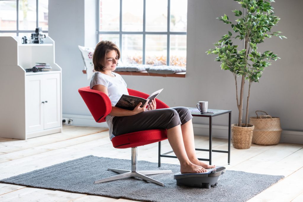 Frau auf einem Sessel mit Zeitschrift und Fußmassagegerät Massage One
